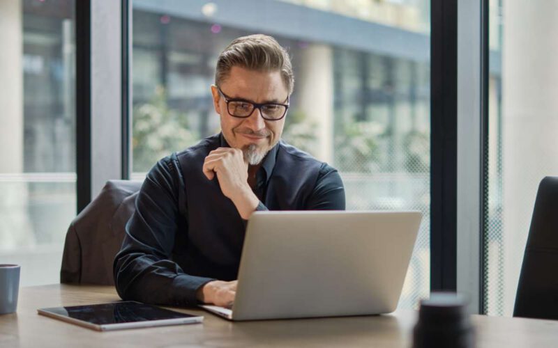 Business portrait - businessman using laptop computer in office, thinking. Happy middle aged man, entrepreneur working online.